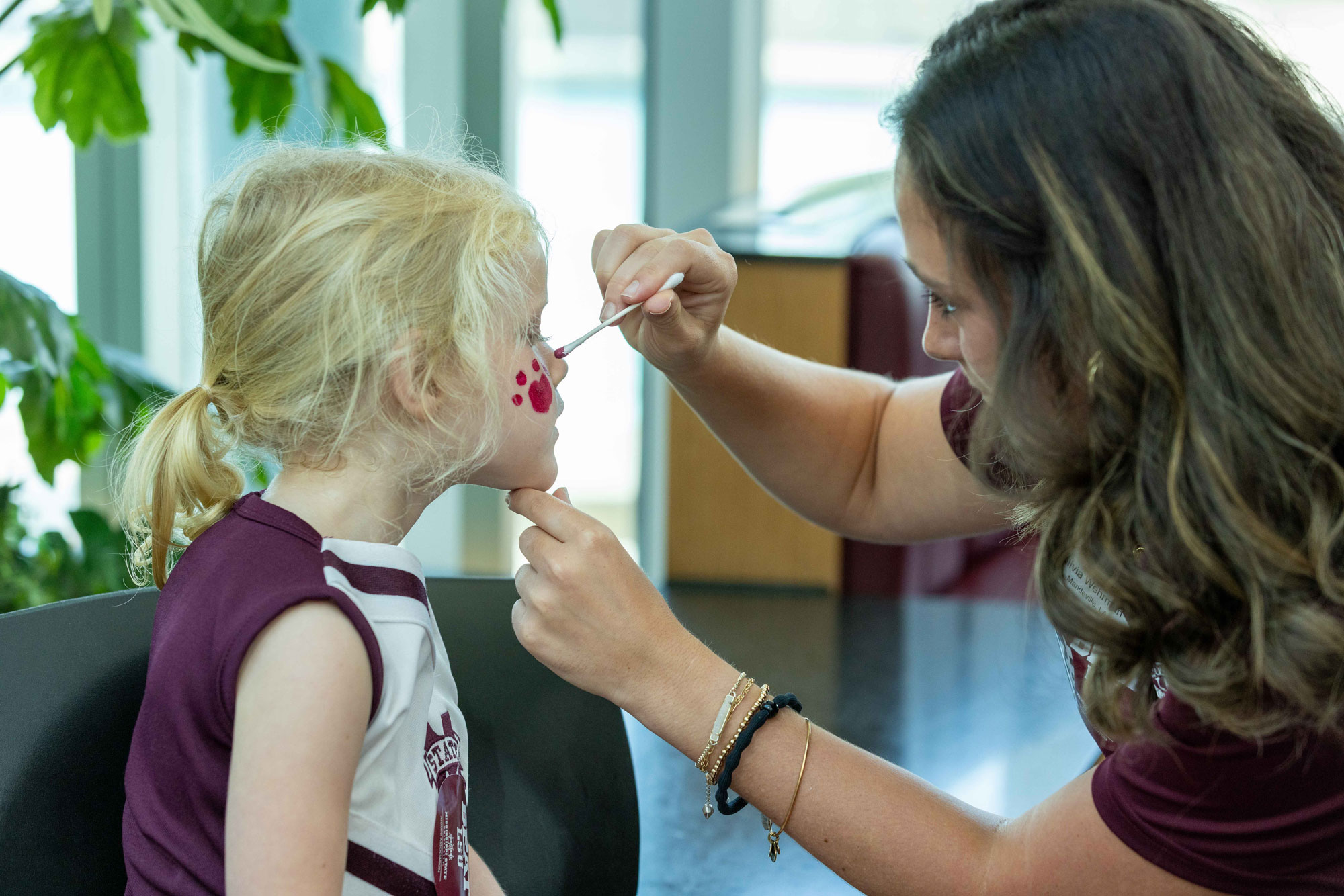 A girl is getting her face painted by a woman in a maroon shirt.