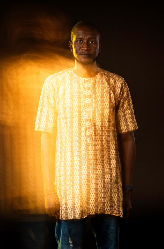 A man in an african shirt standing in front of a dark background.