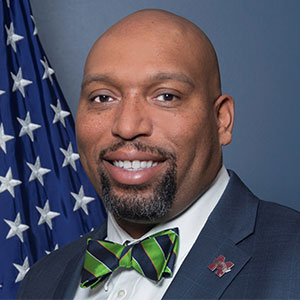 A bald man in a suit and bow tie in front of an american flag.