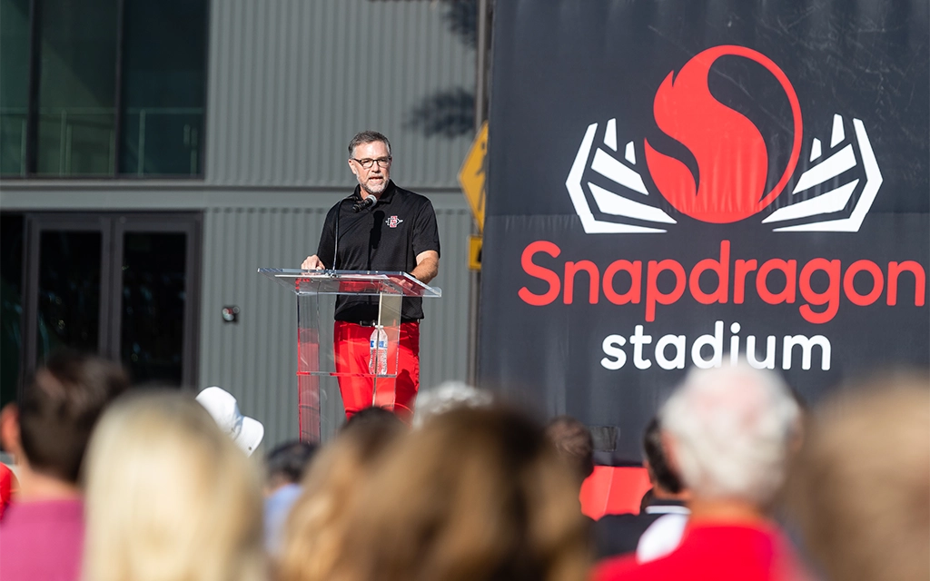 A man speaks at a podium in front of an audience at Snapdragon Stadium. The stadium's logo and name are displayed prominently on a large backdrop behind him.