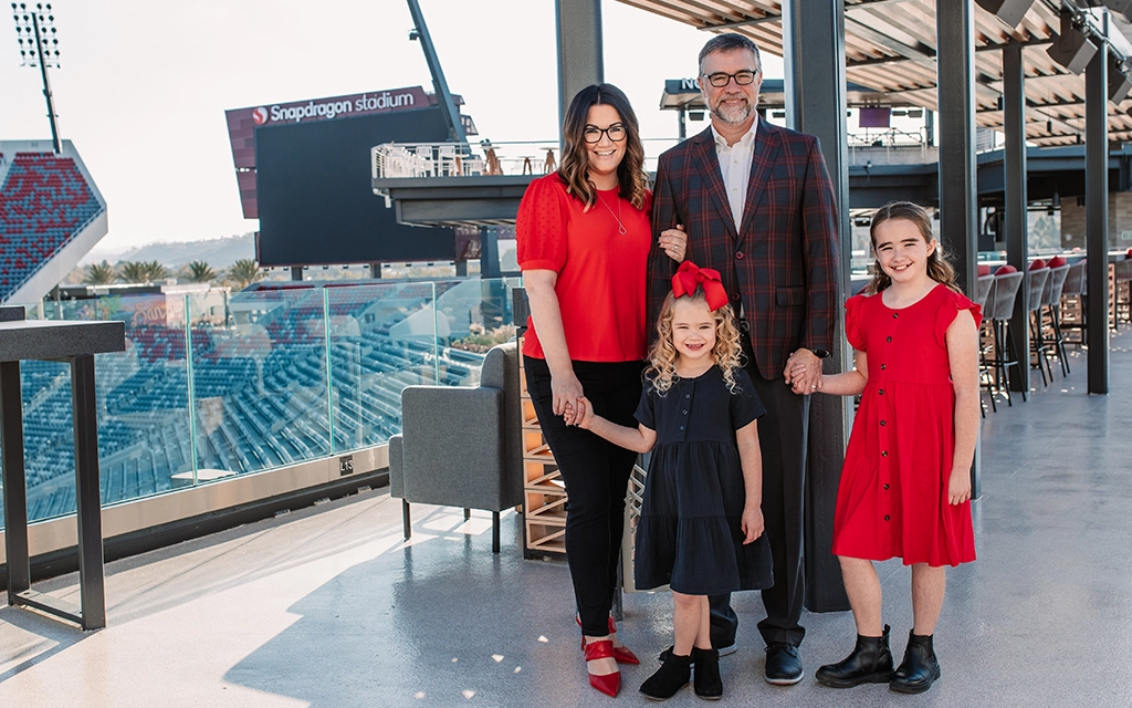 A family of four stands on a terrace overlooking a stadium. The adults and two children are dressed in coordinated red and black outfits.