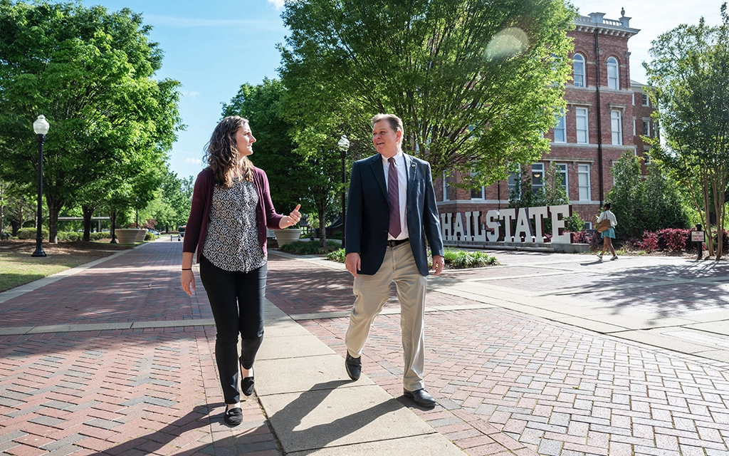 Two people are walking and talking on a brick pathway at a university campus, with a large "CHALL STATE" sign and a red-brick building in the background.