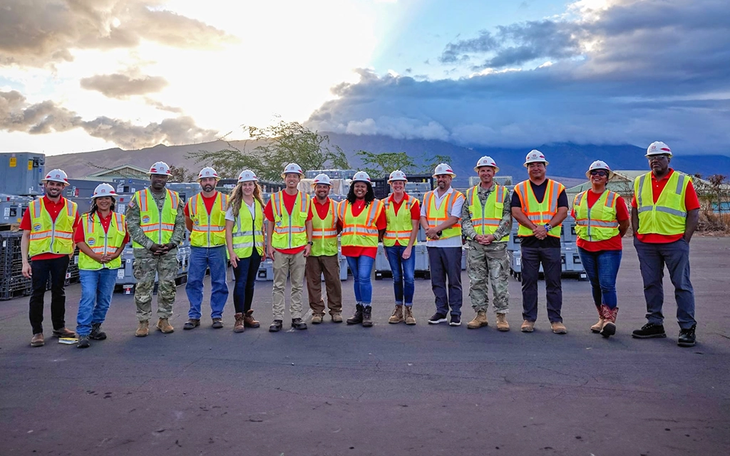 A group of construction workers and engineers wearing hard hats and high-visibility safety vests stand on a paved area with mountains and a partly cloudy sky in the background.