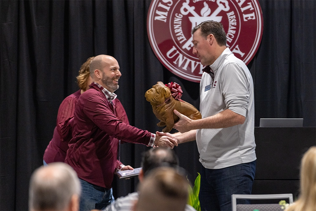 Two men shaking hands; one receives a small statue of a bulldog. A maroon and white university seal is displayed in the background.