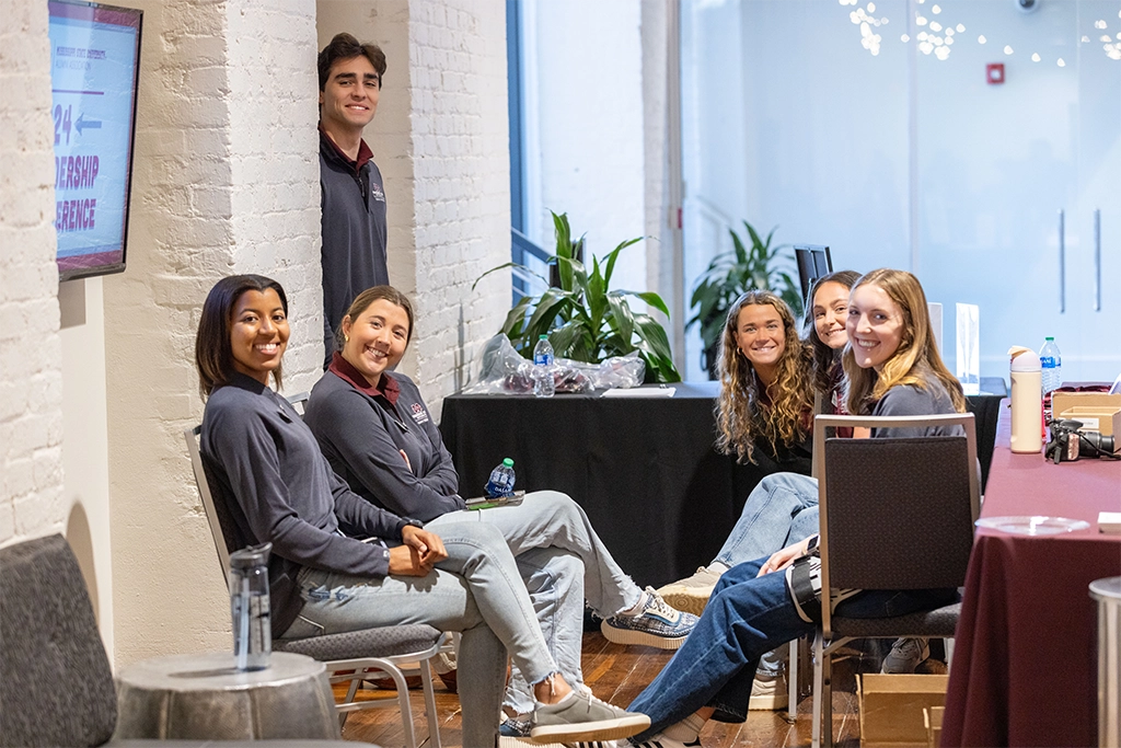 A group of six people are seated and smiling, gathered around a table with various items on it in a well-lit indoor space with white brick walls.