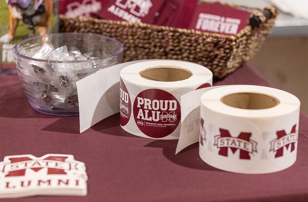 Two rolls of stickers on a table, one with "Proud Alumni" text and the other featuring a logo. A bowl of wrapped candies and a basket with maroon colored items are behind the stickers.