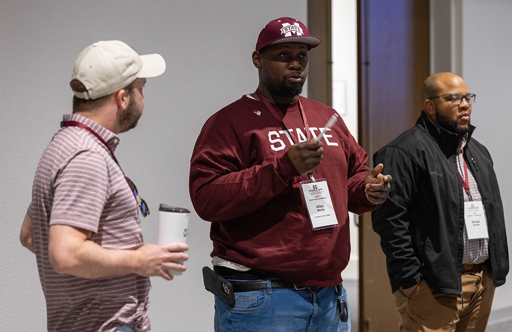 Three men are standing and talking at an indoor event. One man is holding a white cup, another man is speaking, and the third man is looking on. Two of them have visible conference name tags.