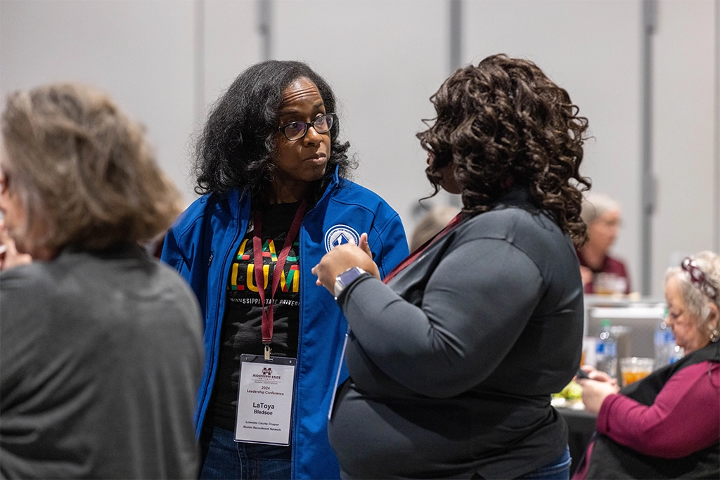 Two people engaged in conversation at an indoor event. One is wearing a blue jacket and name tag, while the other is dressed in a dark top. Several others are seated in the background.