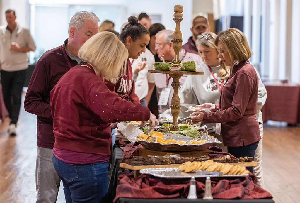 A group of people, wearing maroon and white clothing, serve themselves food from a buffet table arranged with trays of various dishes and a vertical décor piece in the center.