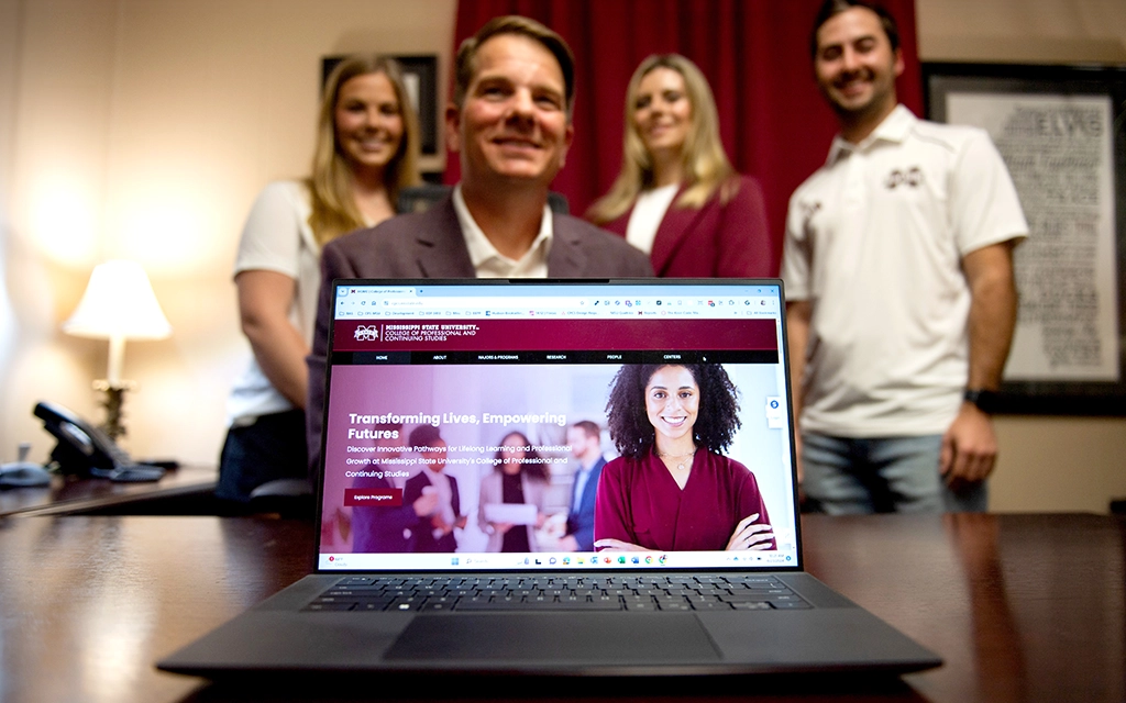 A laptop displaying a university's website with four people, three standing and one seated, smiling in the background in an office setting.