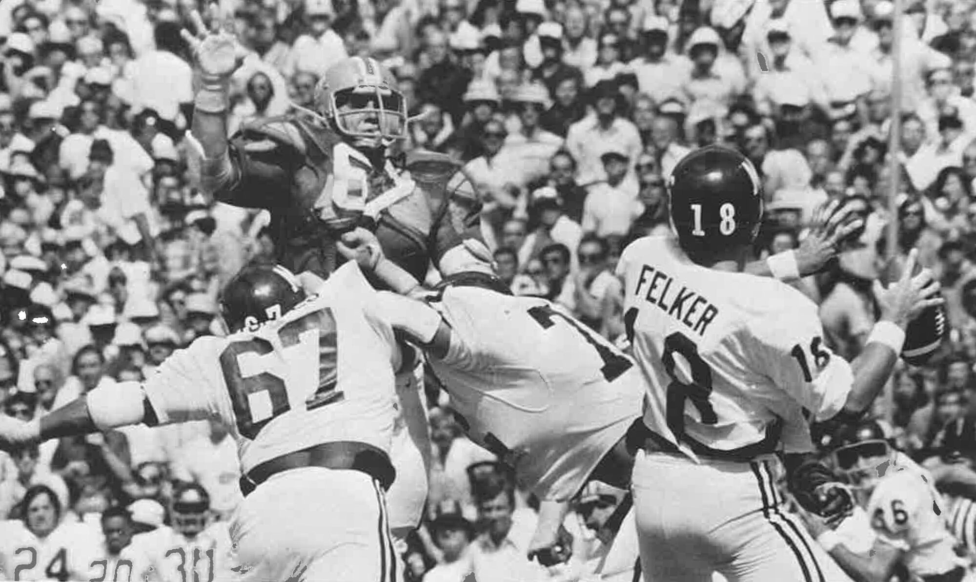 Football game action shot, one player jumping to block a pass as opposing team members, number 16 and 67, attempt to clear a path. Crowd is visible in the background.