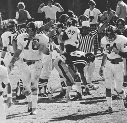 Black and white photo of a football game. Players in white and dark uniforms, referees with striped shirts, and a few onlookers are on the field.