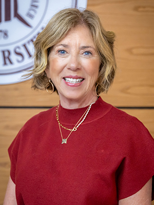 Smiling woman with short blonde hair wearing a red top and gold necklaces stands in front of a wooden wall with a partial university logo in the background.