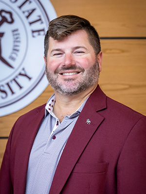 Man in maroon blazer stands smiling in front of a university emblem on a wooden panel wall.