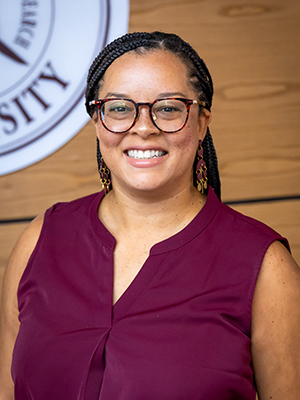 A person with glasses and braided hair smiles, standing in front of a university emblem on a wooden wall.