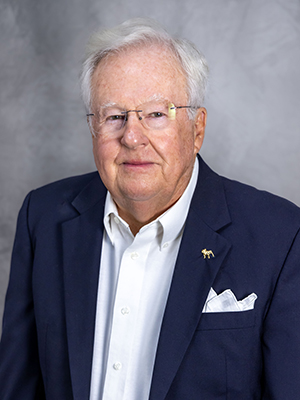 An older man with white hair wearing glasses, a dark suit jacket, and a white shirt, stands against a gray background.