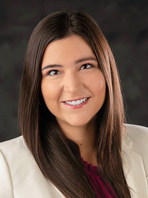 A person with long brown hair and a white blazer smiles in a professional headshot against a dark background.