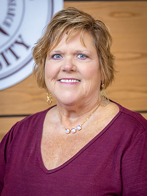 A woman with short hair smiles at the camera, standing in front of a wooden wall with a partial view of a logo.