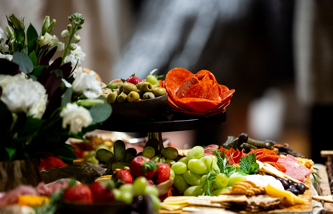 A close-up of a charcuterie board with assorted fruits, meats, and cheeses. A bouquet of flowers is on the left.