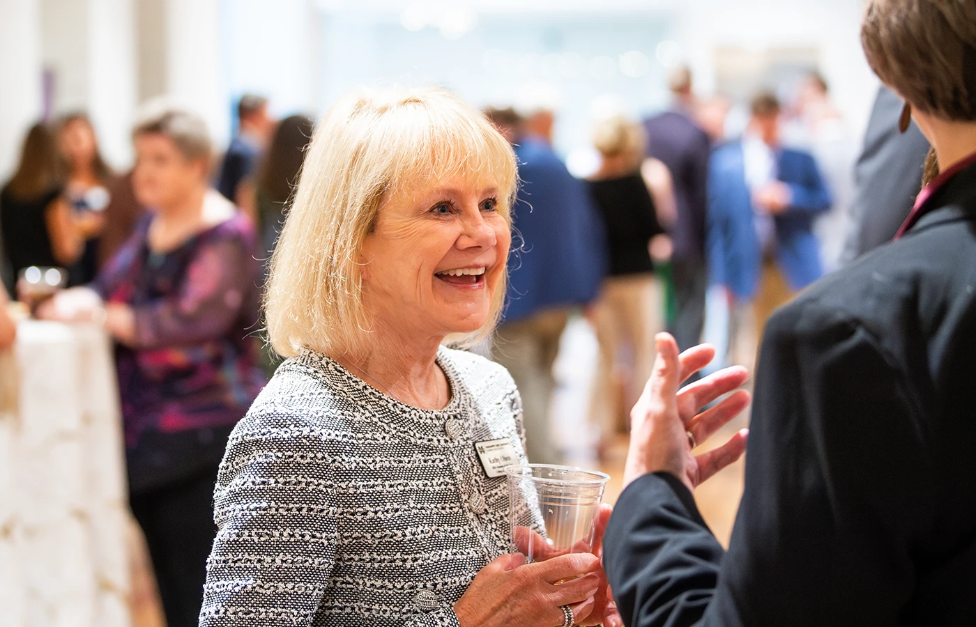 A smiling woman holding a drink engages in conversation at a social event. People are mingling in the background.