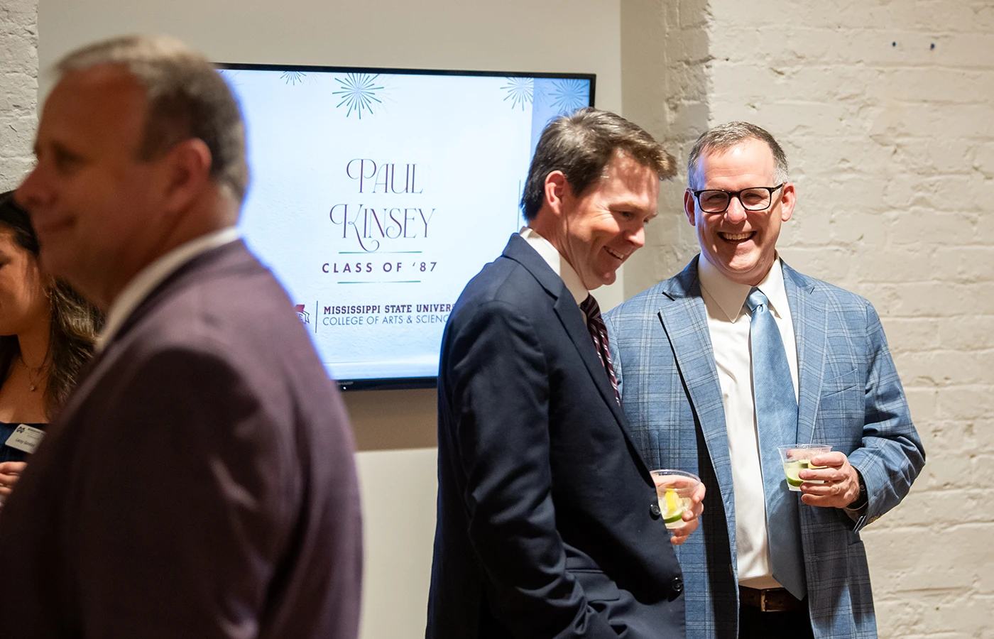 Three men in suits are conversing in a room with a sign in the background that reads "Paul Kinsey Class of '87.