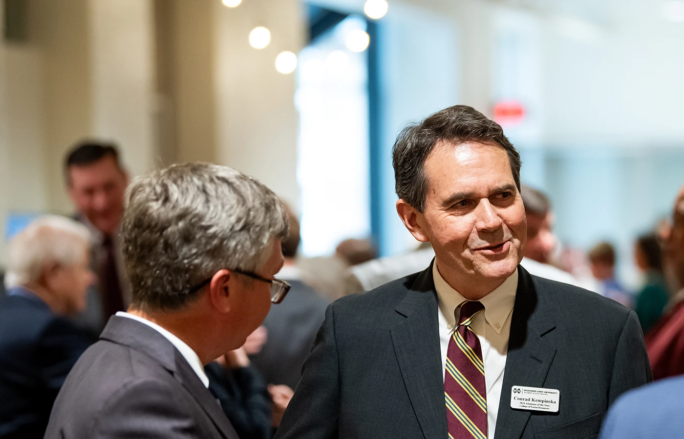 Two men in suits are conversing at a formal event. One wears a name badge. Blurred people and lights are in the background.