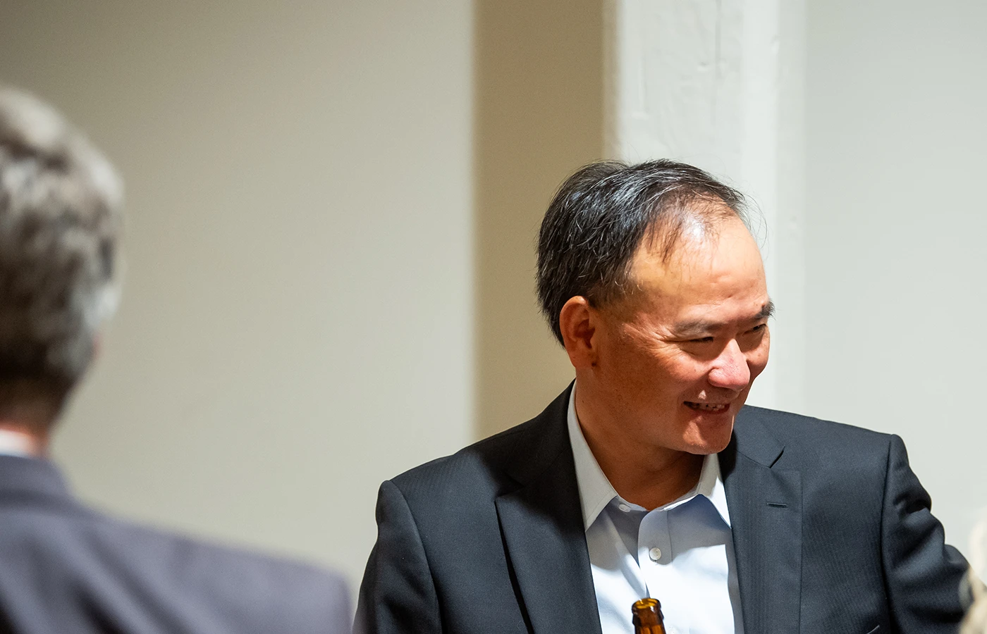 A man in a suit is smiling, standing near a wall, with a blurred person in the foreground.