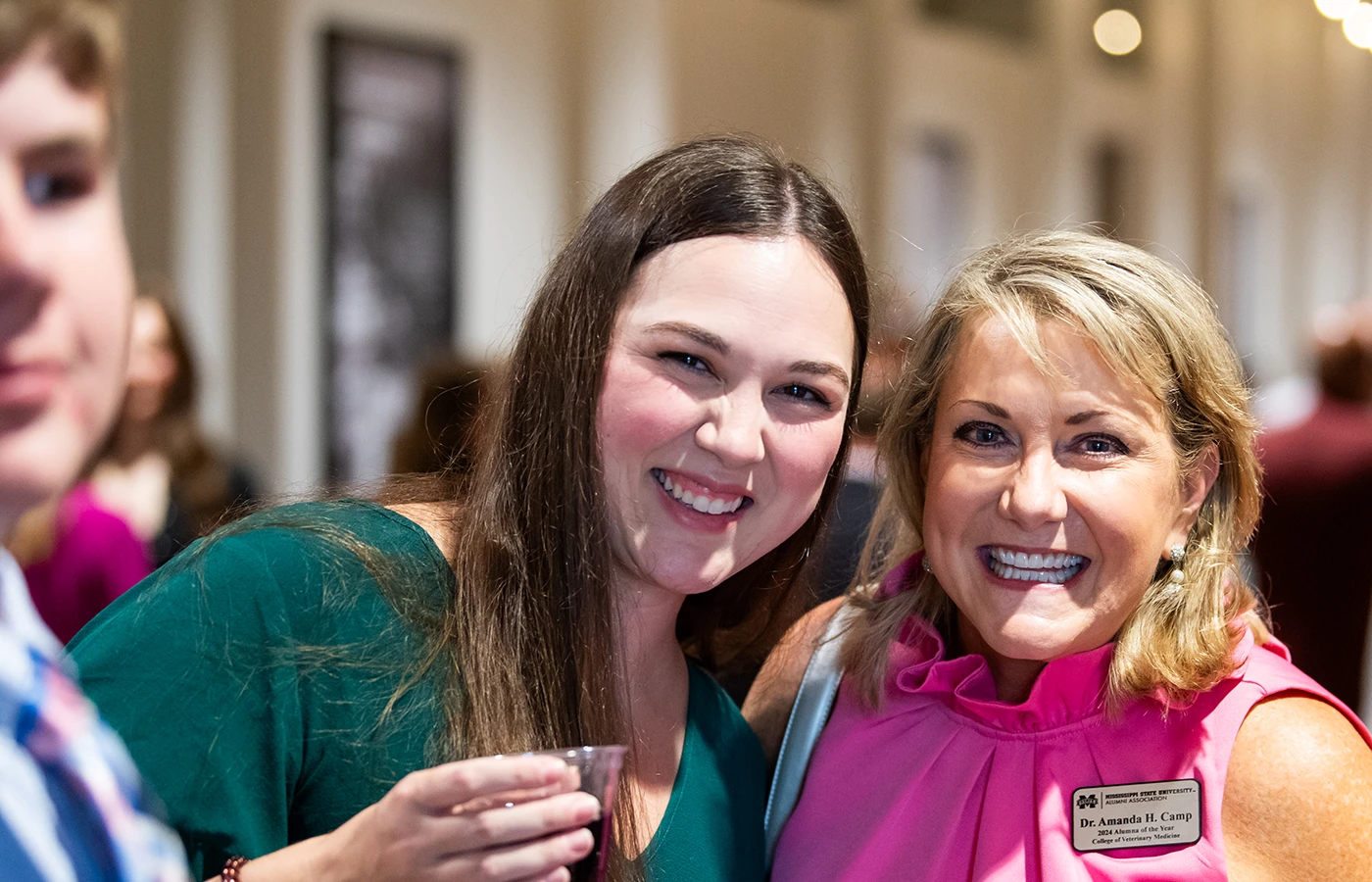 Two women smiling at an event, one holding a drink. The woman on the right wears a name tag.