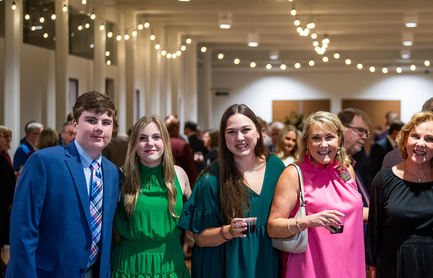 A group of people stands together at an indoor event, with string lights overhead. They appear to be socializing and holding drinks.