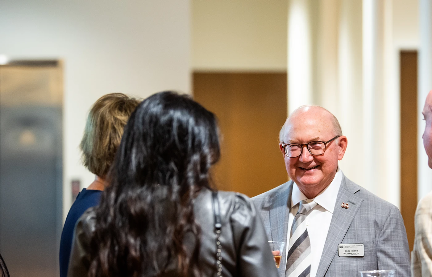 A smiling elderly man in a suit with a name tag talks to a group at an indoor gathering.