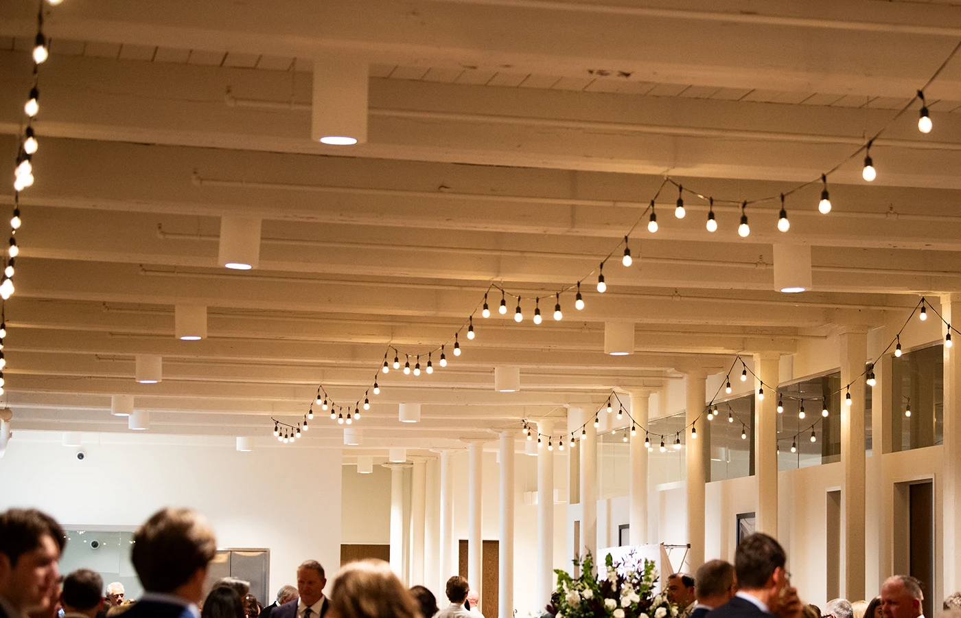 Indoor event space with string lights hanging from the ceiling and people mingling below.