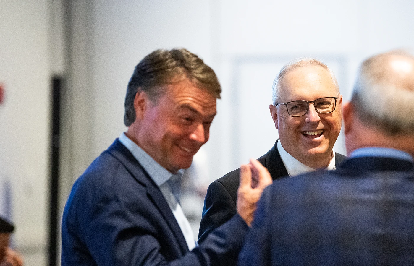 Three men in business attire are engaged in conversation, with two smiling and the focus on the man in the foreground gesturing.