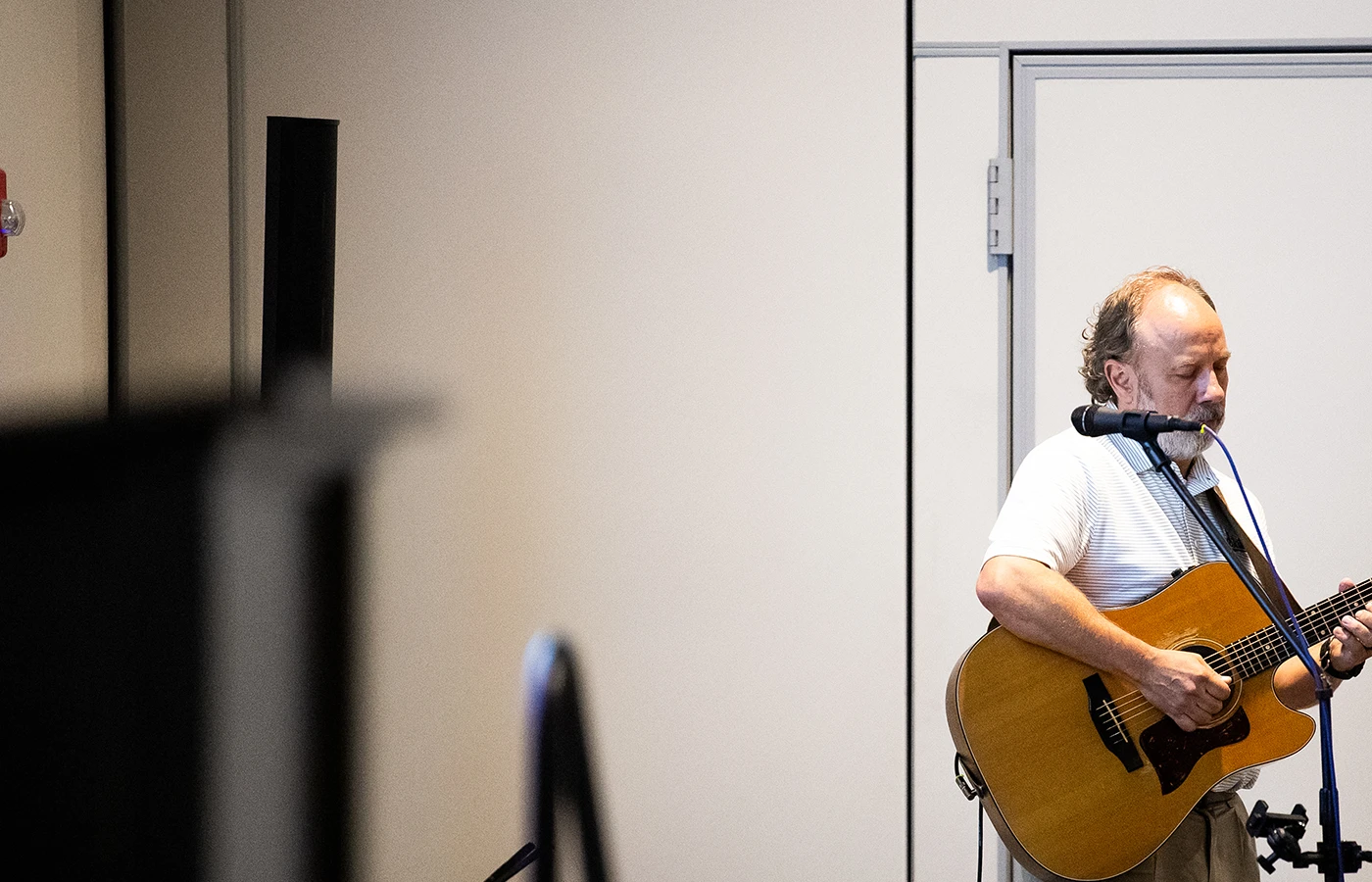 A man stands playing an acoustic guitar near a microphone against a plain wall.