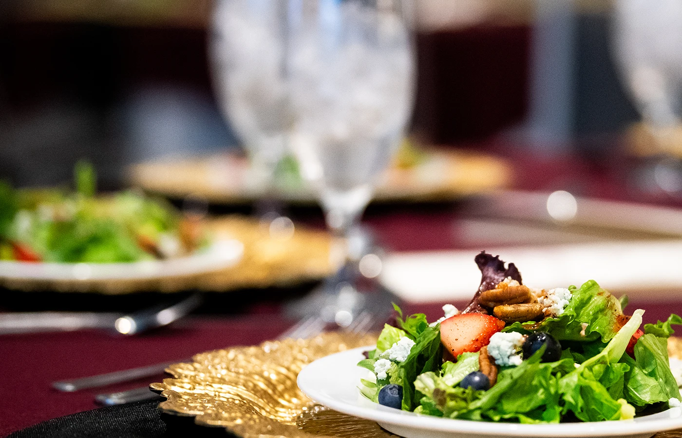 A close-up of a plated salad with mixed greens, strawberries, pecans, and crumbled cheese. Ice water glasses and more salad plates are in the background.