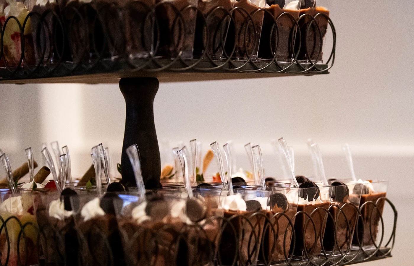 A two-tiered stand holds an assortment of dessert cups, each with clear spoons. Some desserts are topped with berries, chocolate, and cream.