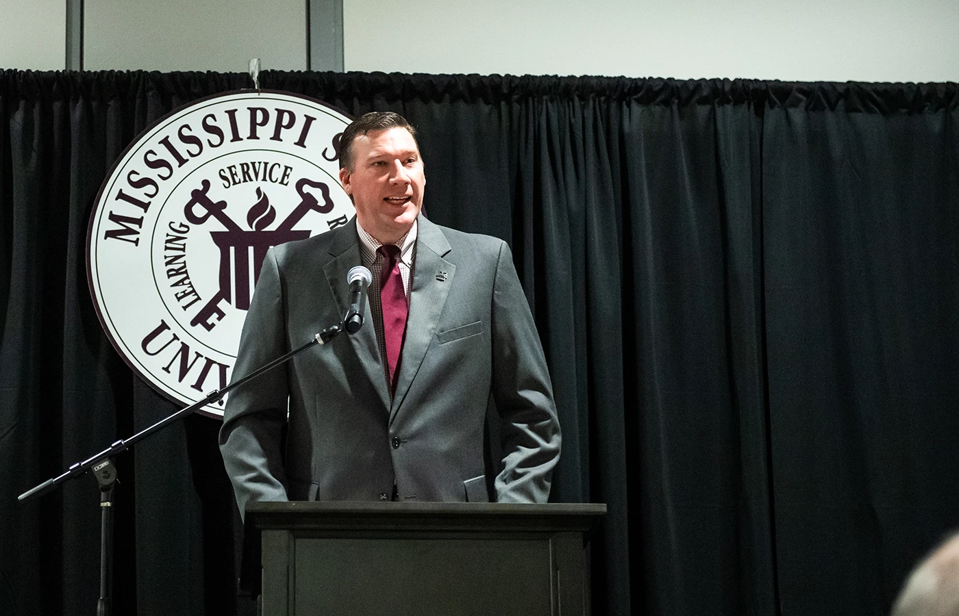 A man in a suit speaks at a podium with a Mississippi State University emblem in the background.