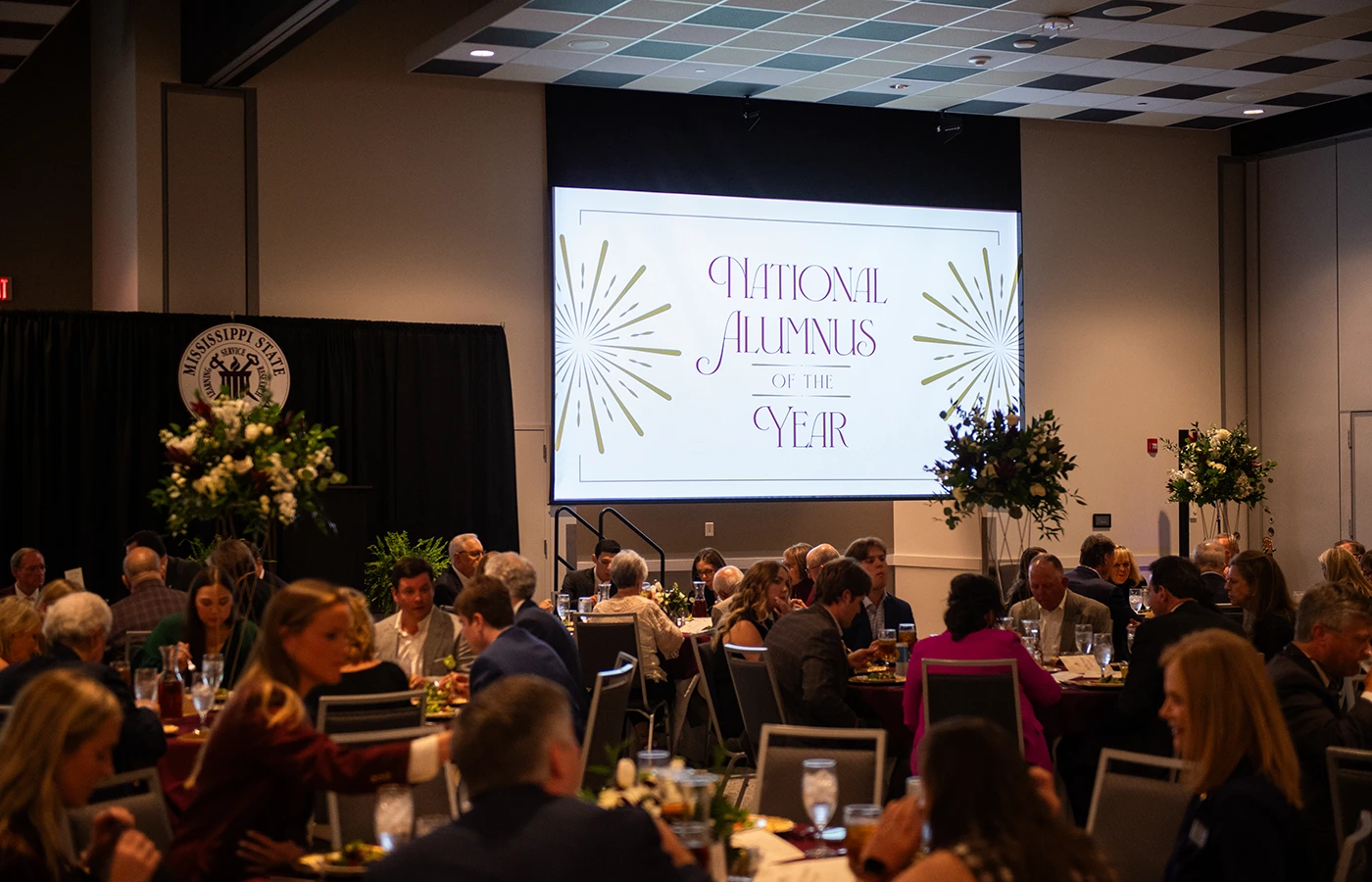 A conference room with attendees seated at tables, a large screen displaying "National Alumnus of the Year," and flower arrangements near the stage.