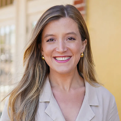 Smiling person with long hair wearing a beige jacket stands in front of a yellow wall.