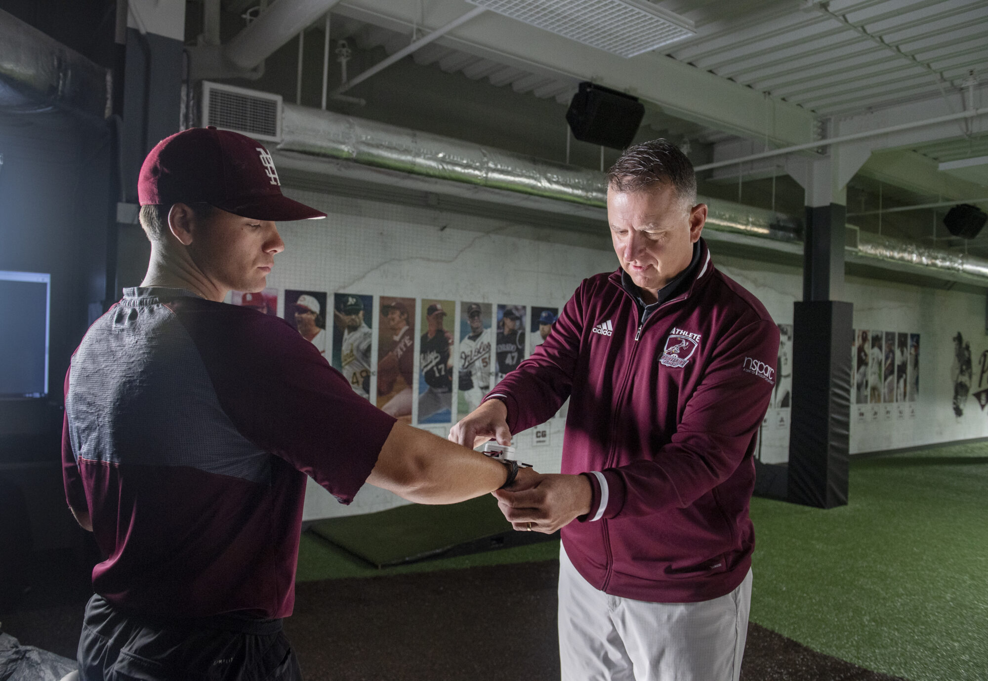 A coach adjusts the wrist of a baseball player in an indoor training facility. Both are wearing maroon sports attire. Posters of baseball players line the wall in the background.