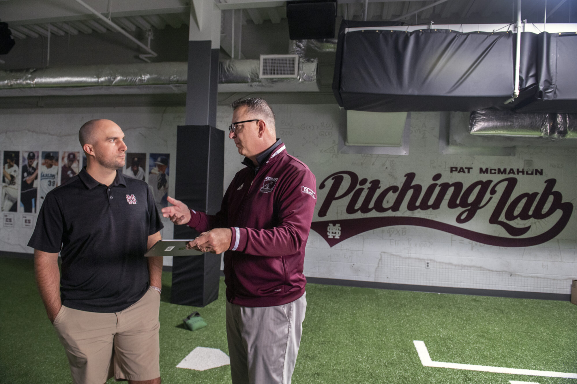 Two men stand inside a training facility called the "Pitching Lab". One is holding a tablet and listening while the other gestures as he speaks. The room has artificial turf flooring.