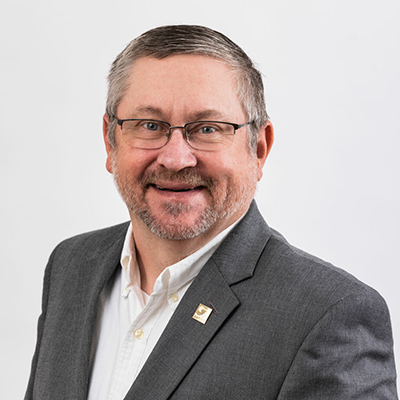 A man with glasses, a beard, and a mustache smiles while wearing a gray suit jacket and a white shirt.