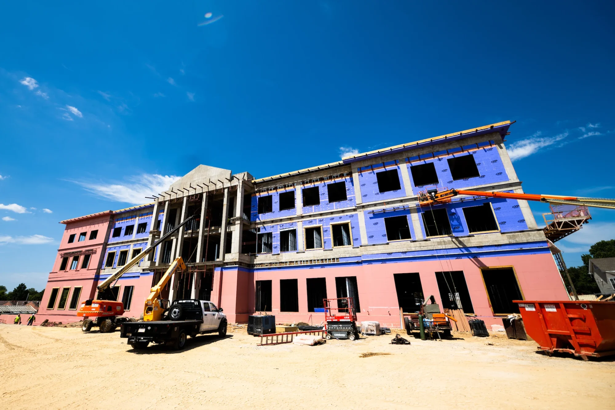 A large, multi-story building under construction with two cherry pickers and various construction equipment on a sandy site under a clear blue sky.