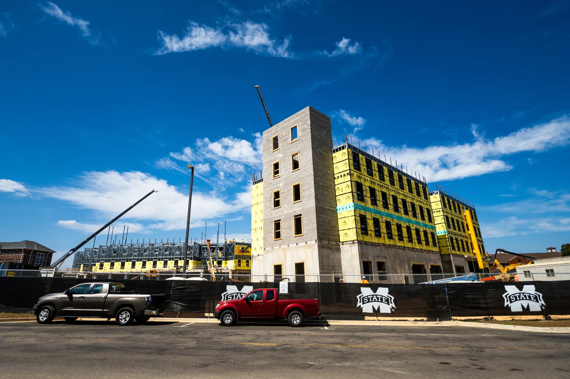 Construction site with a multi-story building under development, surrounded by fencing. Two trucks are parked in the foreground under a clear blue sky.