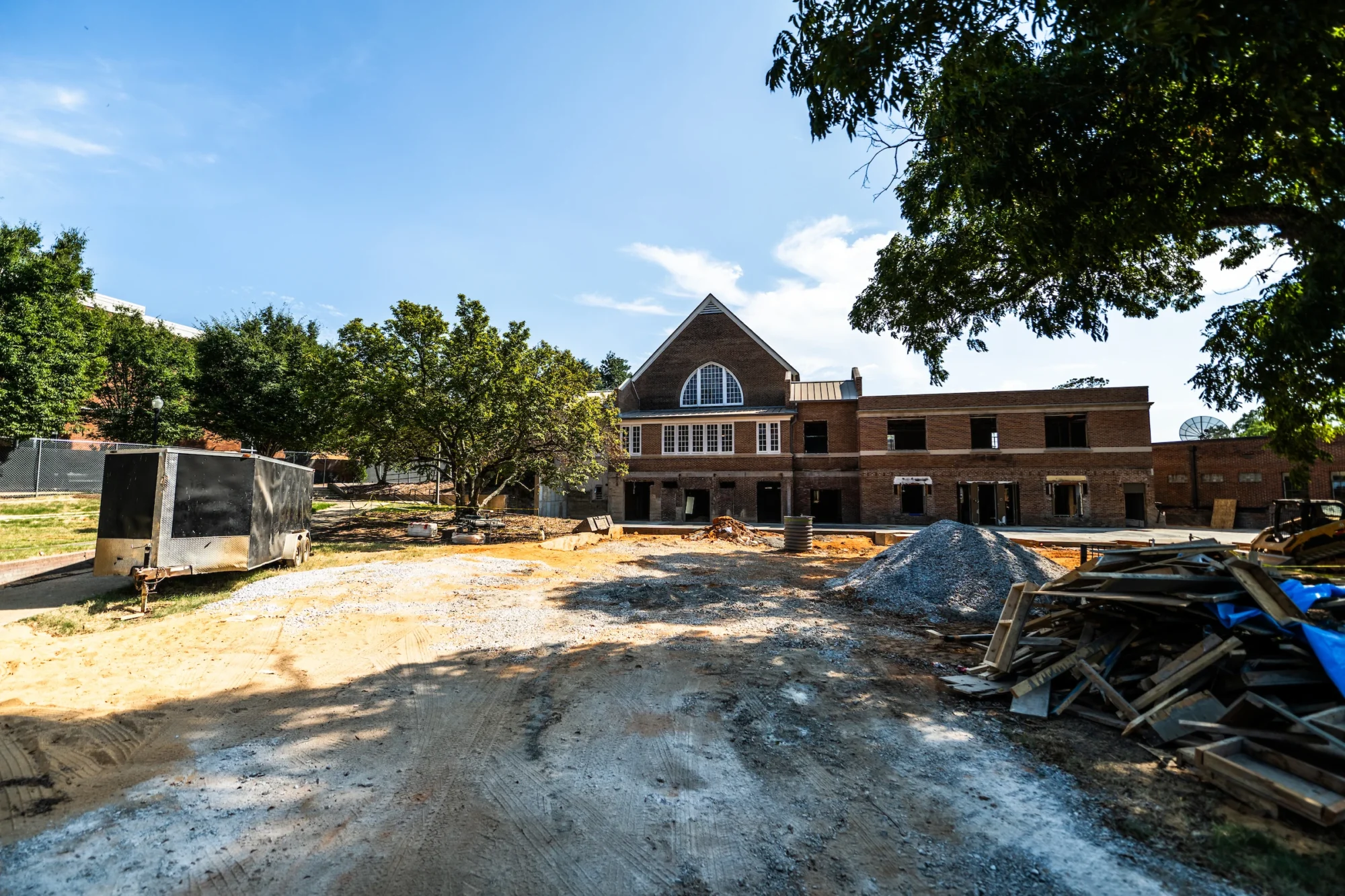 A construction site with a partially built brick building, surrounded by trees, dirt, and building materials under a clear sky.