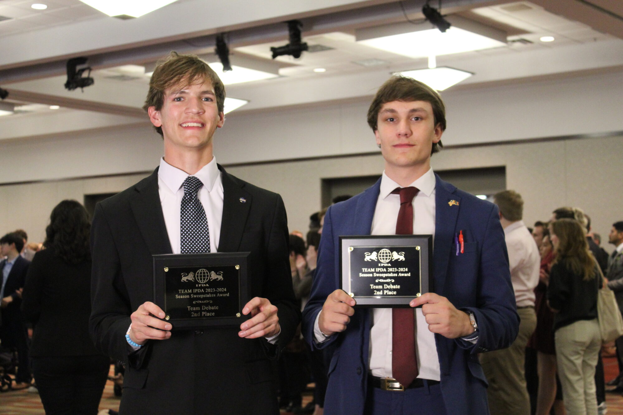 Two young men in suits hold award plaques at an indoor event. A crowd is visible in the background.