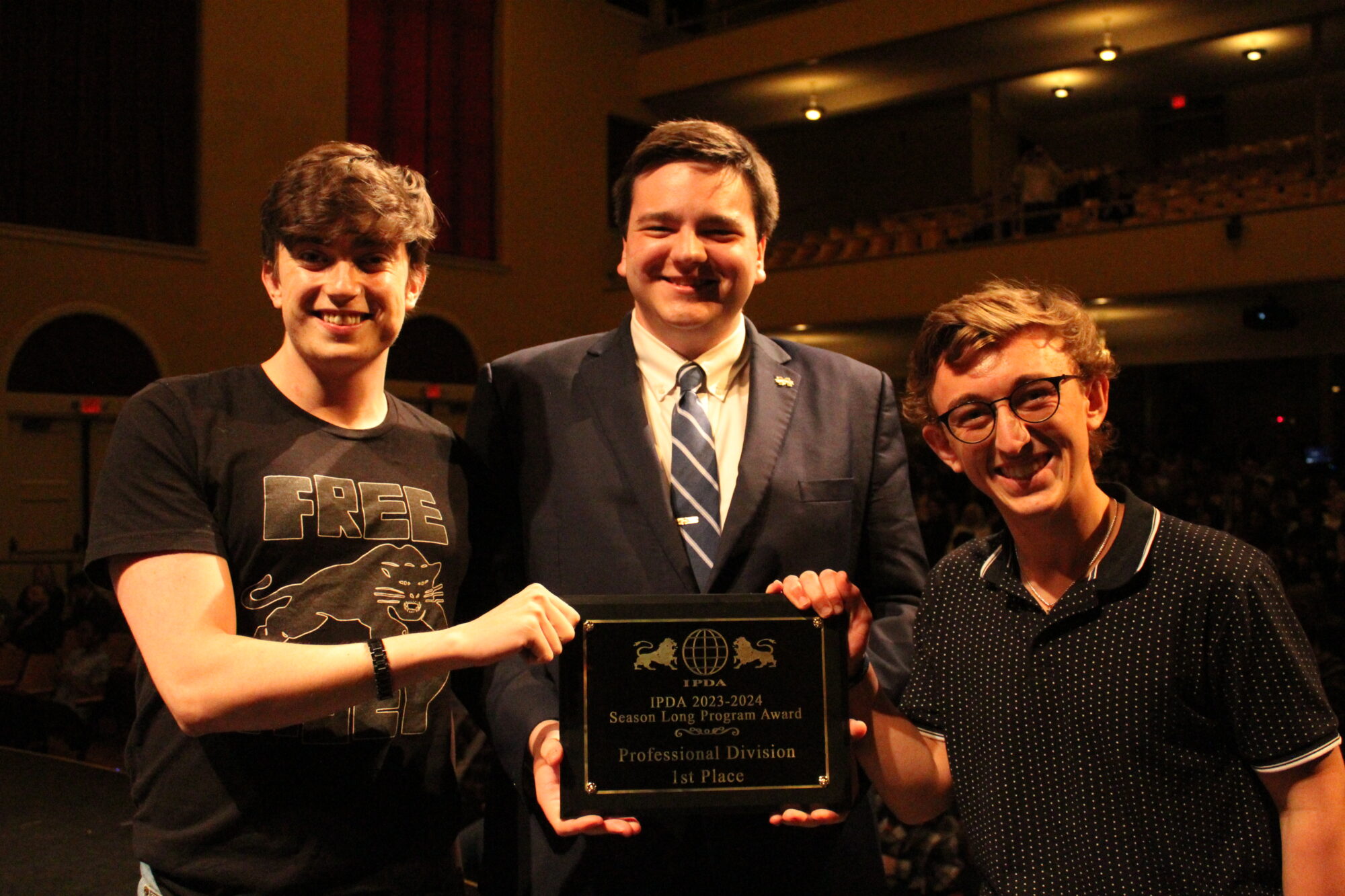 Three people stand on stage, smiling and holding a plaque that reads "IPDA 2023-2024 Season Long Program Award, Champions, Professional Division, 1st Place.