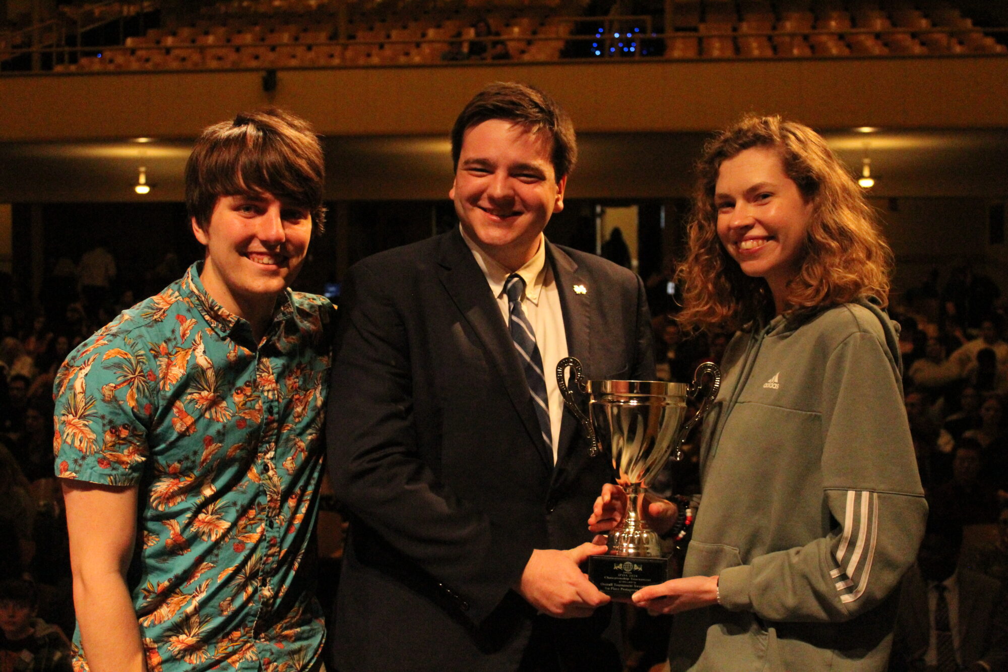 Three people pose with a trophy in an auditorium. The person in the center is wearing a suit, while the others wear casual clothing.