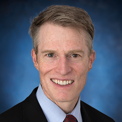 A man in a suit and tie smiles against a blue background.