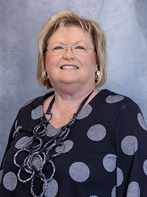 A woman with short blonde hair smiles while wearing glasses, a black top with large gray polka dots, and a necklace.