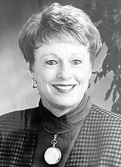 A grayscale portrait of a smiling person with short hair, wearing a checkered blazer and pendant necklace.